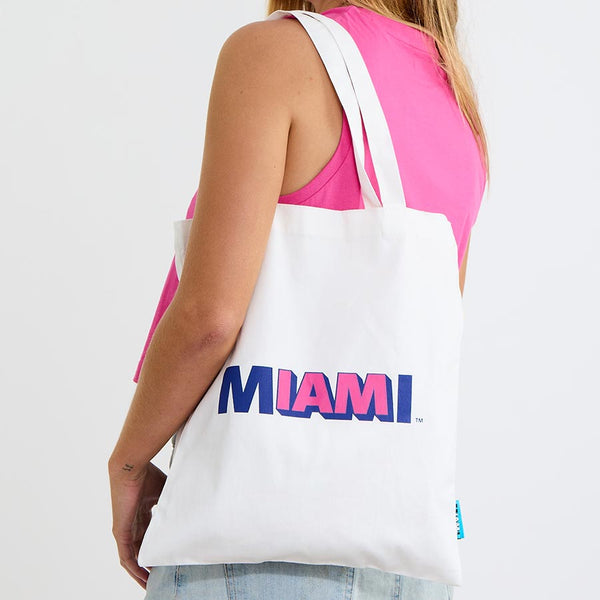 Woman carrying the white Coral Miami tote bag over her shoulder, I AM MIAMI logo on the front